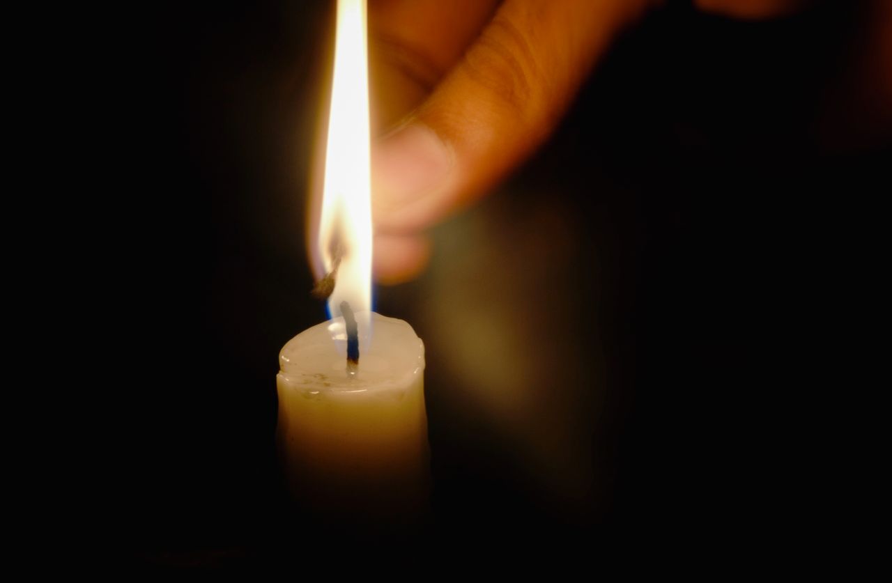 A close-up of a candle lit in darkness, with a hand visible near the flame.